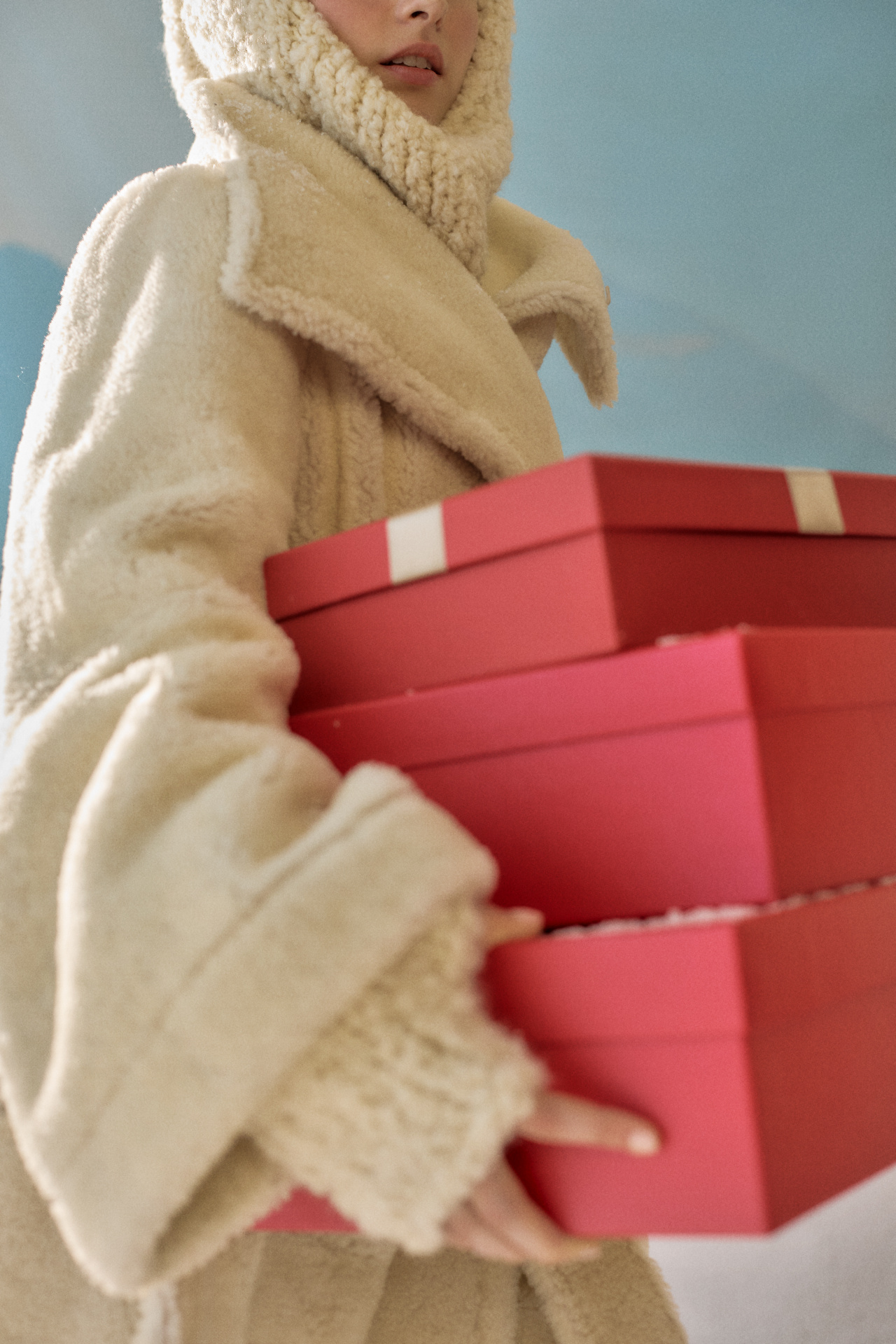 Model in cream shearling coat carrying stacked red holiday gift boxes