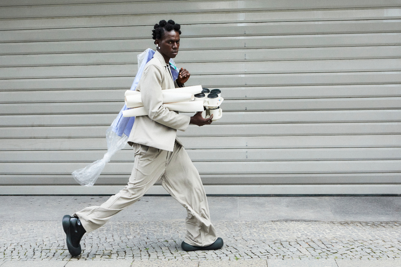 Man in a beige suit walking briskly with coffees and posters